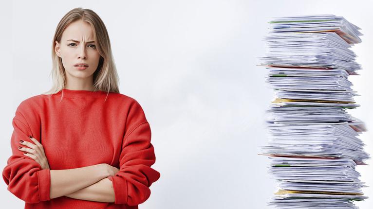 Eine junge Frau steht neben einem hohen Stapel ungeöffneter Briefe und sieht den Betrachtenden verärgert an