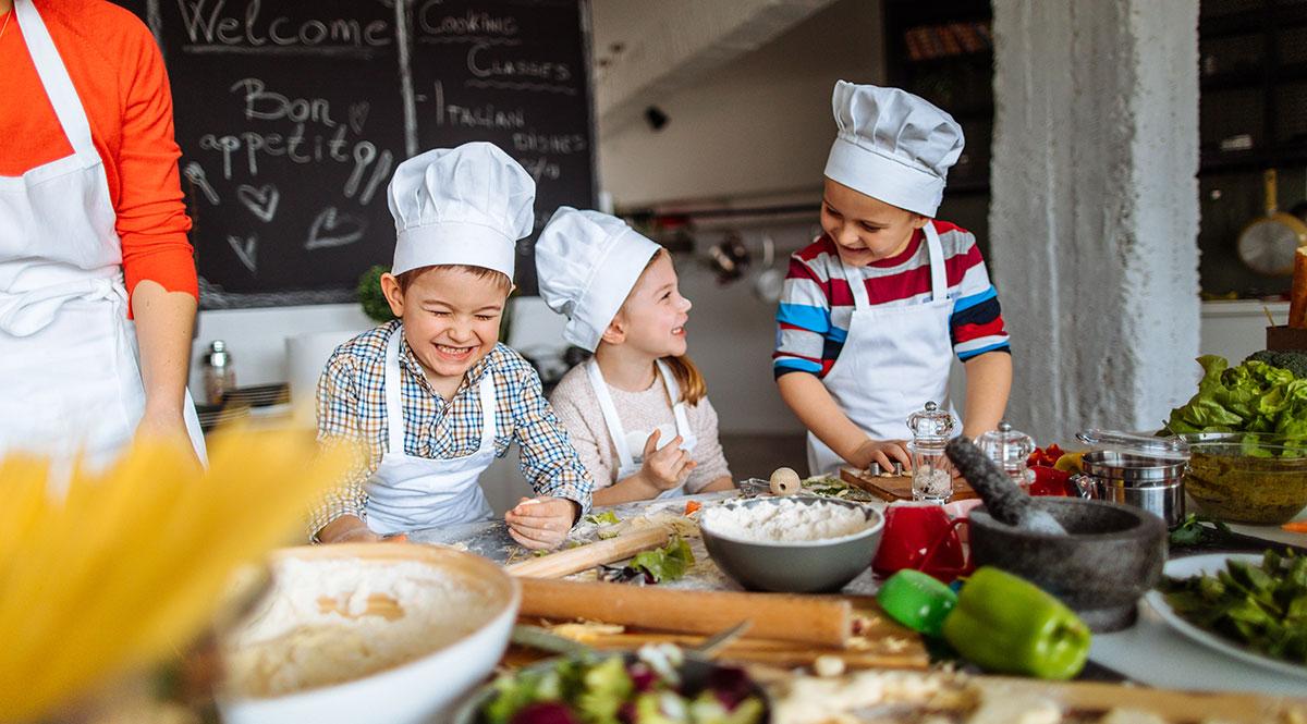 Drei Kinder mit Kochmützen und Schürzen lachen bei der Essenzubereitung