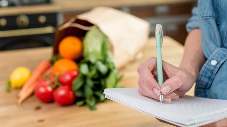 Jemand schreibt in ein Notizbuch, in Hintergrund liegt auf einem Tisch eine Tragetasche mit Gemüse