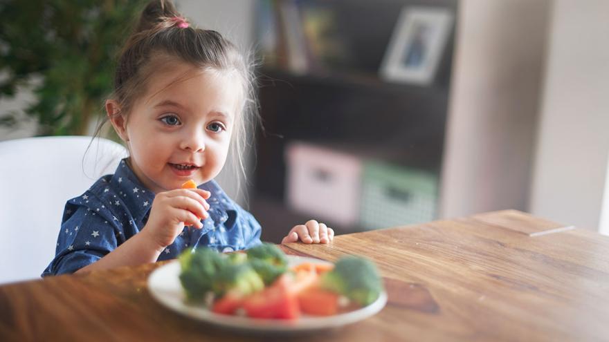 Ein Mädchen sitzt am Tisch und isst mit der Hand verschiedene Gemüse von einem Teller 