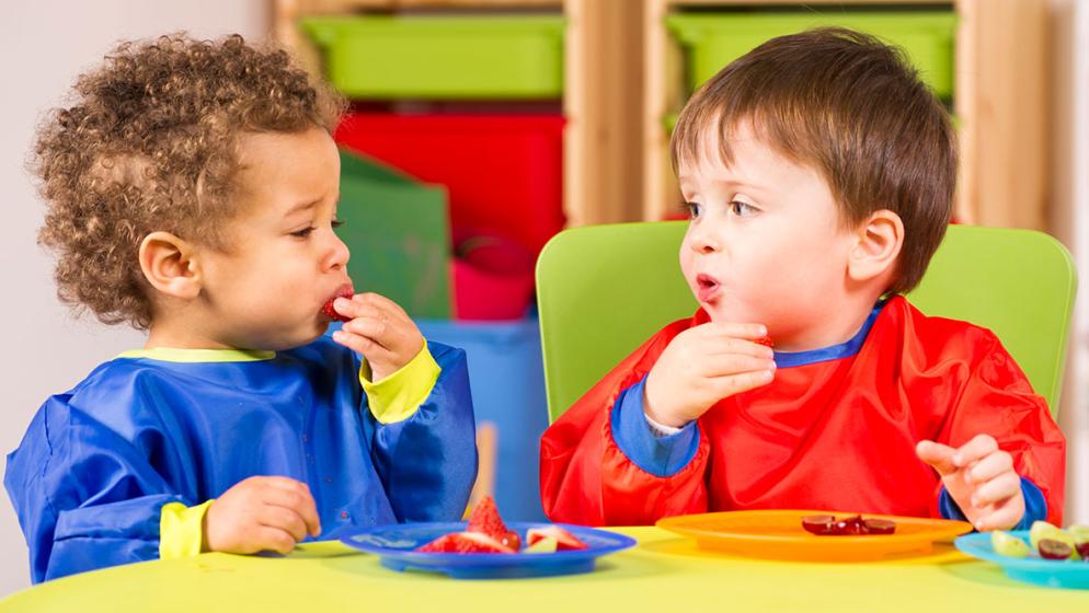 Zwei Kleinkinder mit Lätzchen essen Obst von Tellern 